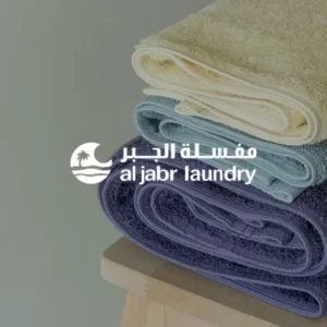 Stack of neatly folded towels on a wooden stool with Al Jabr Laundry logo in Arabic and English.